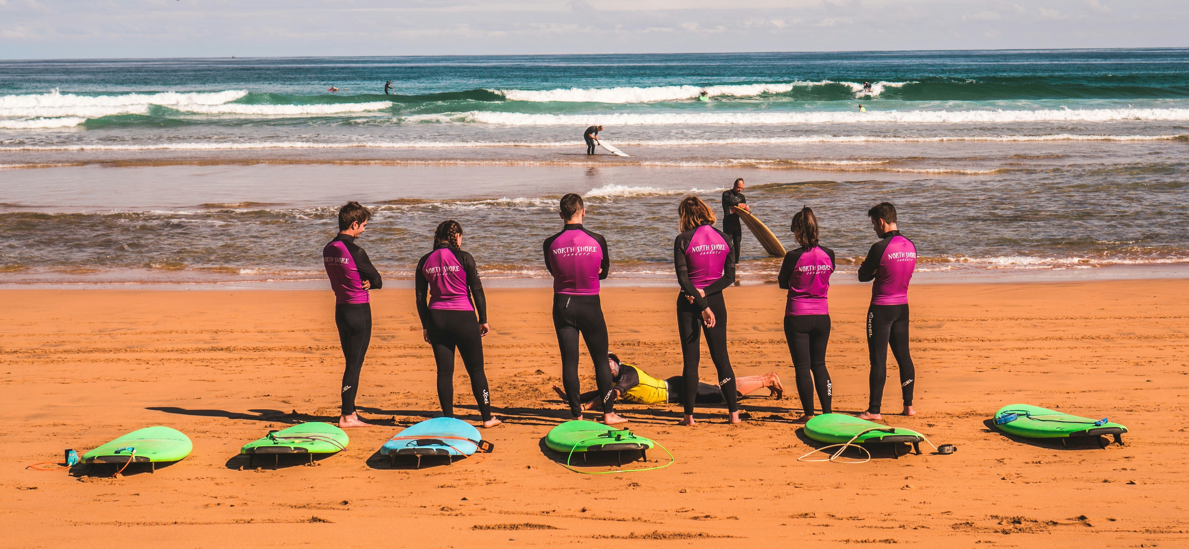 Group of surf students in wetsuits on a sandy beach with surfboards, watching waves roll in